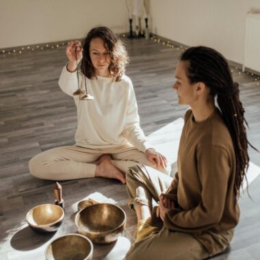 Two individuals engage in a meditation session with Tibetan singing bowls in a peaceful, sunlit room.